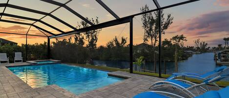 A swimming pool with a glass enclosure, blue lounge chairs, and a view of a river at sunset.