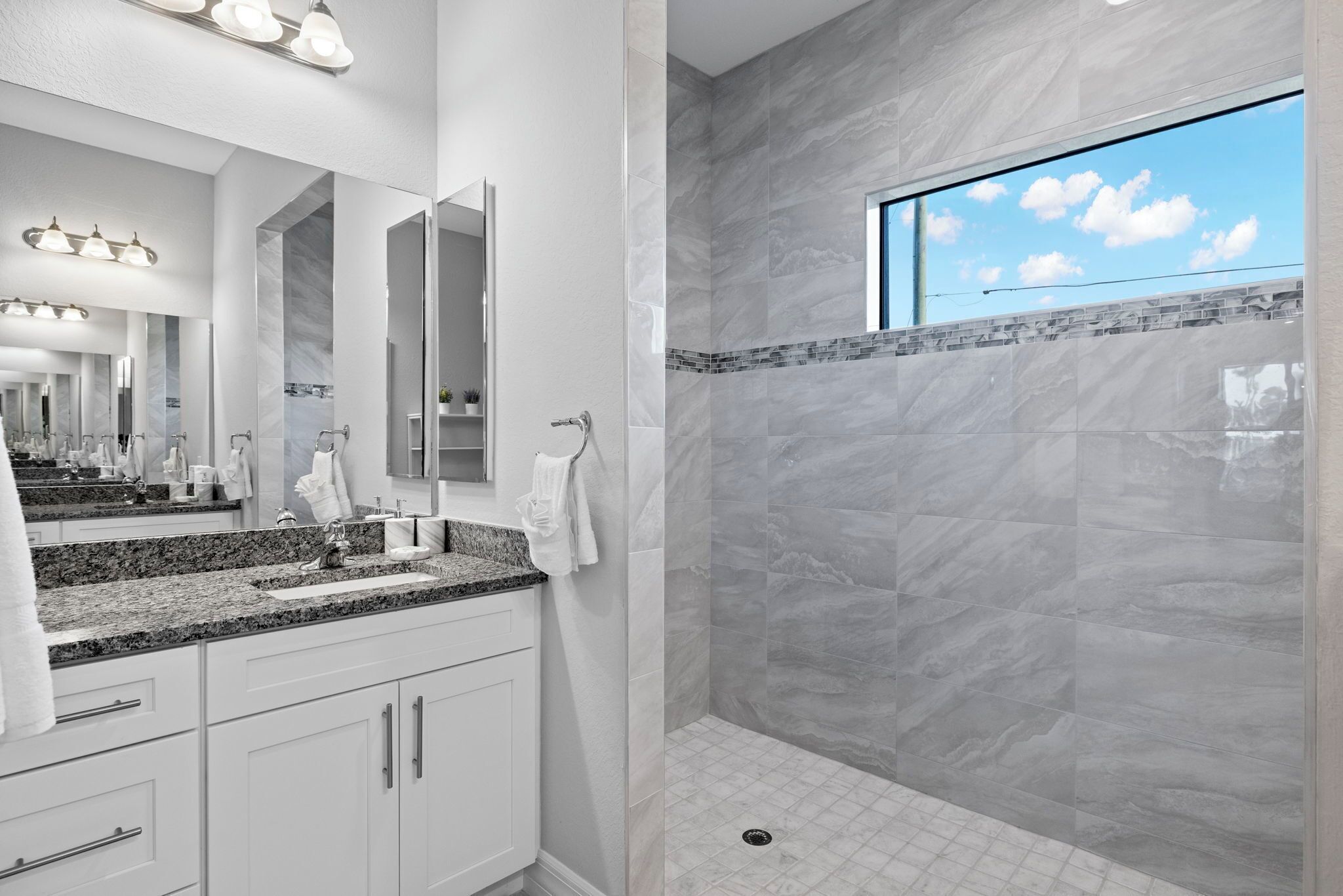 A modern bathroom with a large mirror, granite countertop, and a shower with a view of the sky.