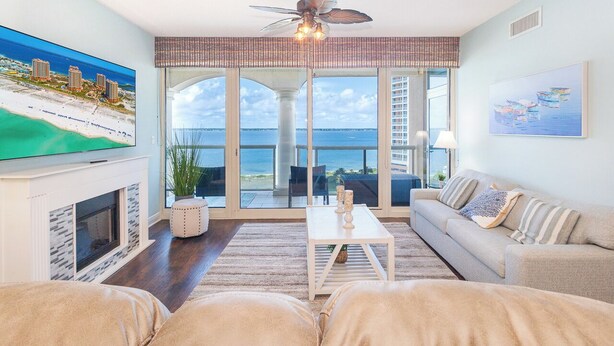 A living room with a large TV, a fireplace, a ceiling fan, a sofa, and a view of the ocean through large glass doors. - New Listing P5-0906 2B Portofino Island View! (Pensacola Beach)