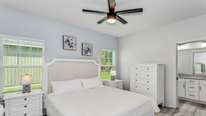 Un dormitorio con cama, mesitas de noche, cómoda, ventilador de techo y dos ventanas con persianas.