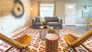 A living room with a grey sofa, a coffee table, and two wooden chairs. There is a patterned rug on the floor, a ceiling fan, and a door with a framed picture on the wall.