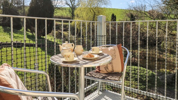 Ein Metalltisch mit Teekanne, Tassen und einem Teller Essen, auf einem Balkon mit Blick auf eine grüne Landschaft und Bäume.