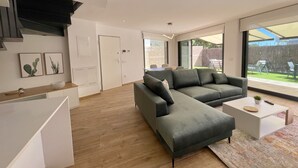 Un salon moderne avec un canapé d’angle, une table basse et un coin repas comprenant une table et des chaises. Une porte-fenêtre coulissante donne accès à un patio aménagé avec du mobilier d’extérieur.