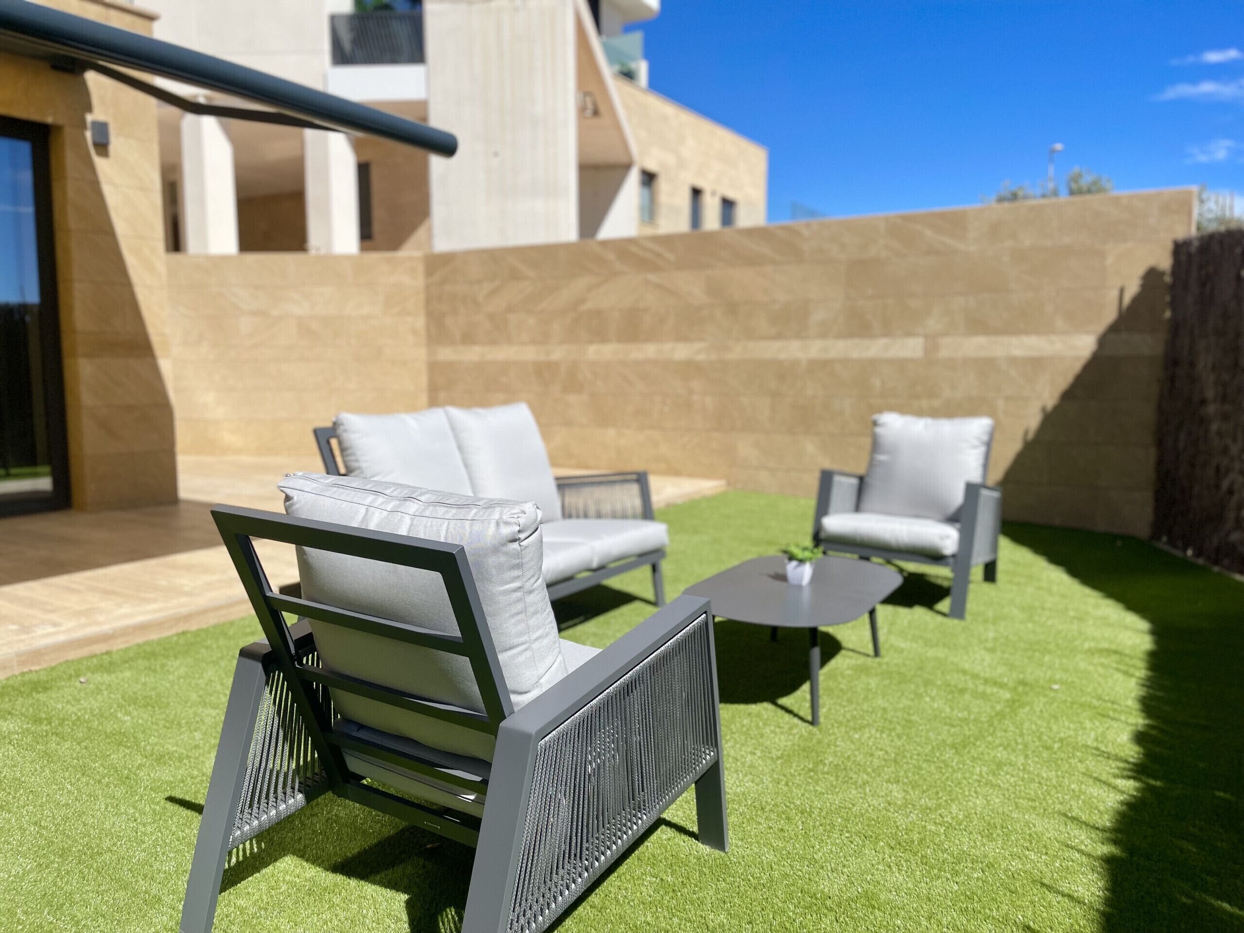 Un espacio exterior moderno con zona de estar, sofá, sillones, una mesita y una estructura de pérgola.