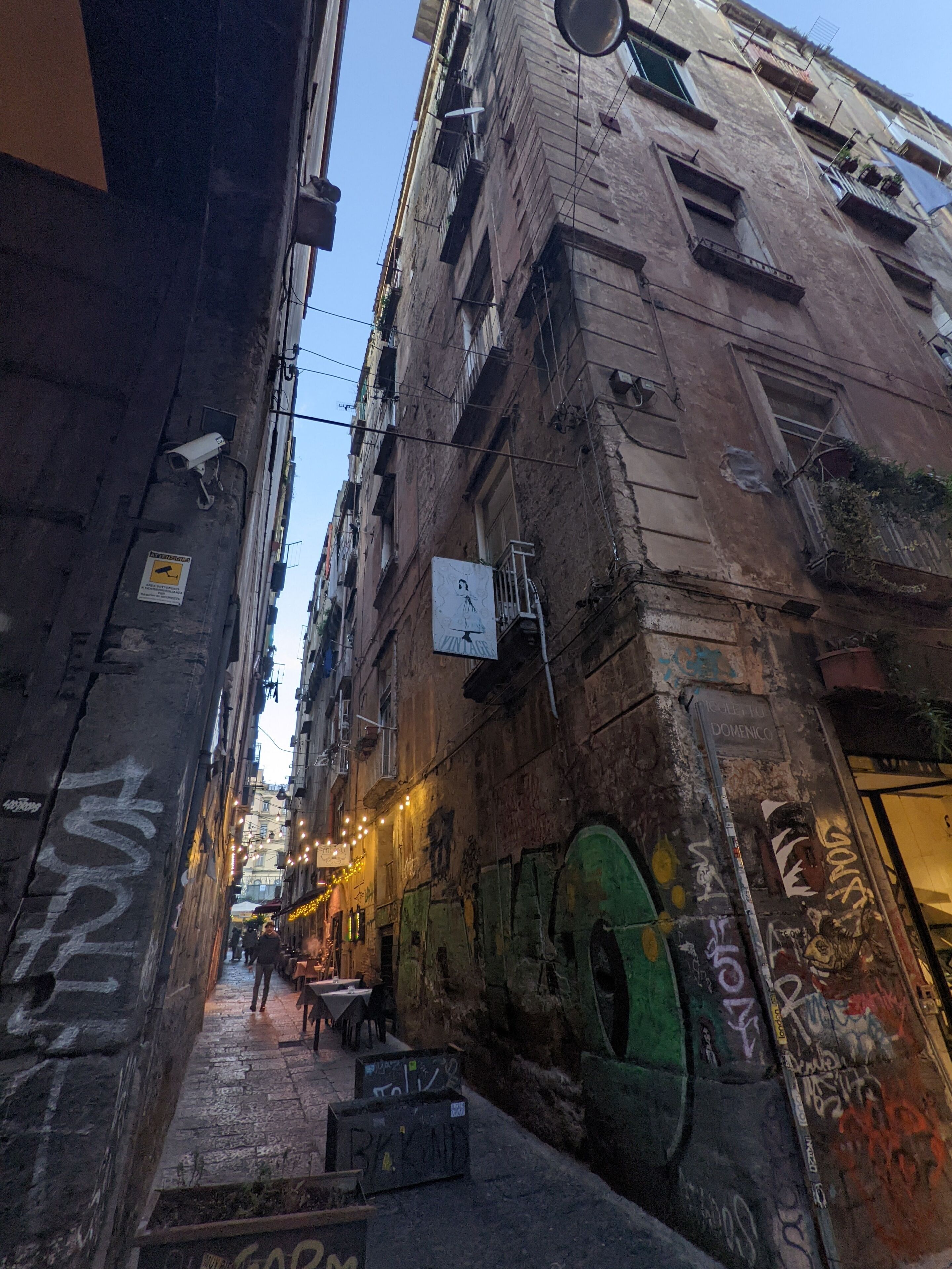 A narrow alleyway with graffiti on the walls, a small outdoor seating area with tables and chairs, and a building with multiple windows and balconies.
