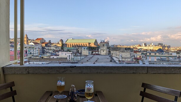 A table with two glasses of beer and a decorative item, overlooking a cityscape with historic buildings and a church. - Napoli City Panorama (Naples)