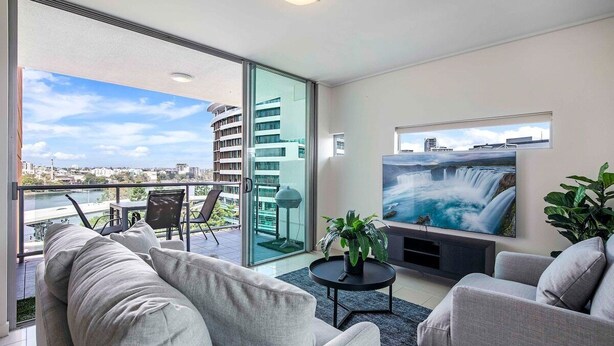 Un salon moderne doté d’un grand téléviseur, de sièges confortables, d’un balcon offrant une vue sur la ville et d’une porte vitrée donnant sur un espace extérieur.