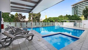 Une piscine avec des chaises longues, un espace abrité et une vue sur un bâtiment moderne en arrière-plan.