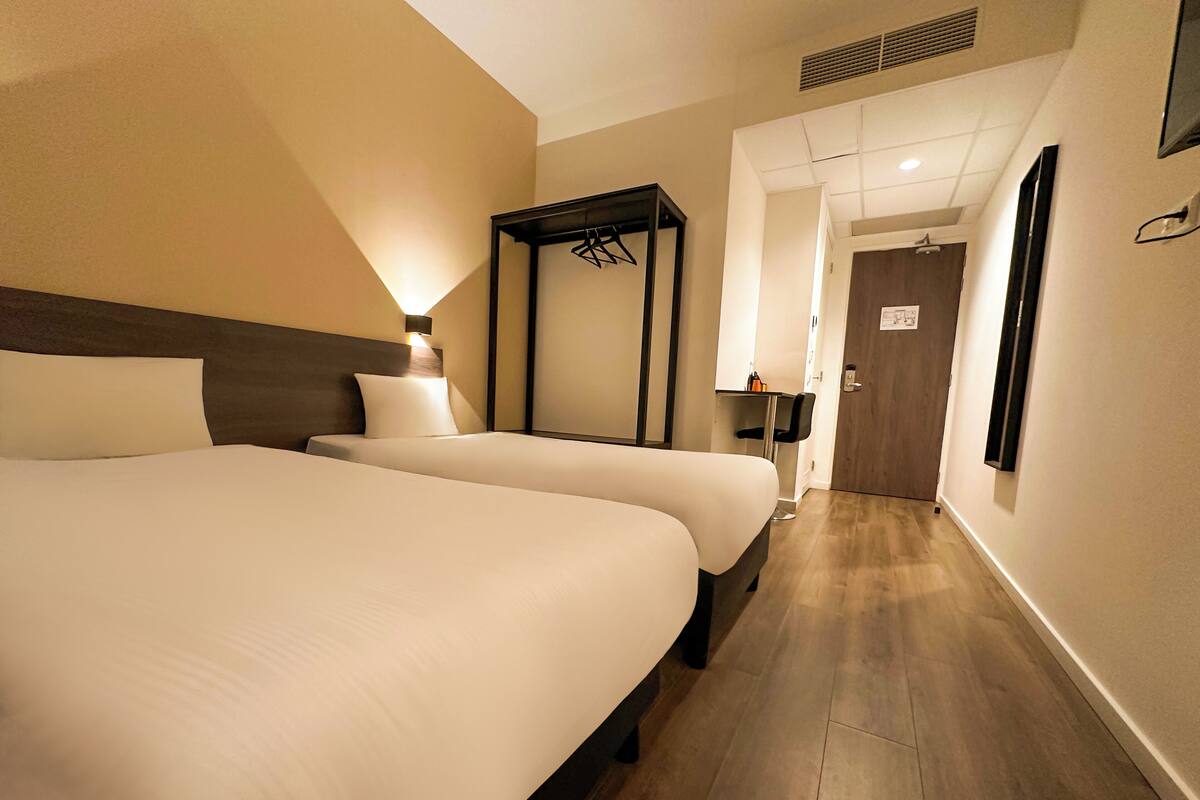 A hotel room with two beds, a bedside table, a wardrobe, and a mirror.