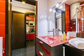 Ein modernes Badezimmer mit einem roten Waschbecken, einem beleuchteten Spiegel, einer Duschkabine und einer sichtbaren Küche im Hintergrund.