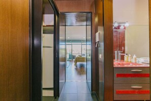 A hotel corridor leading to a room with a bed, a bathroom with a sink and toiletries, and a view of the outdoors.