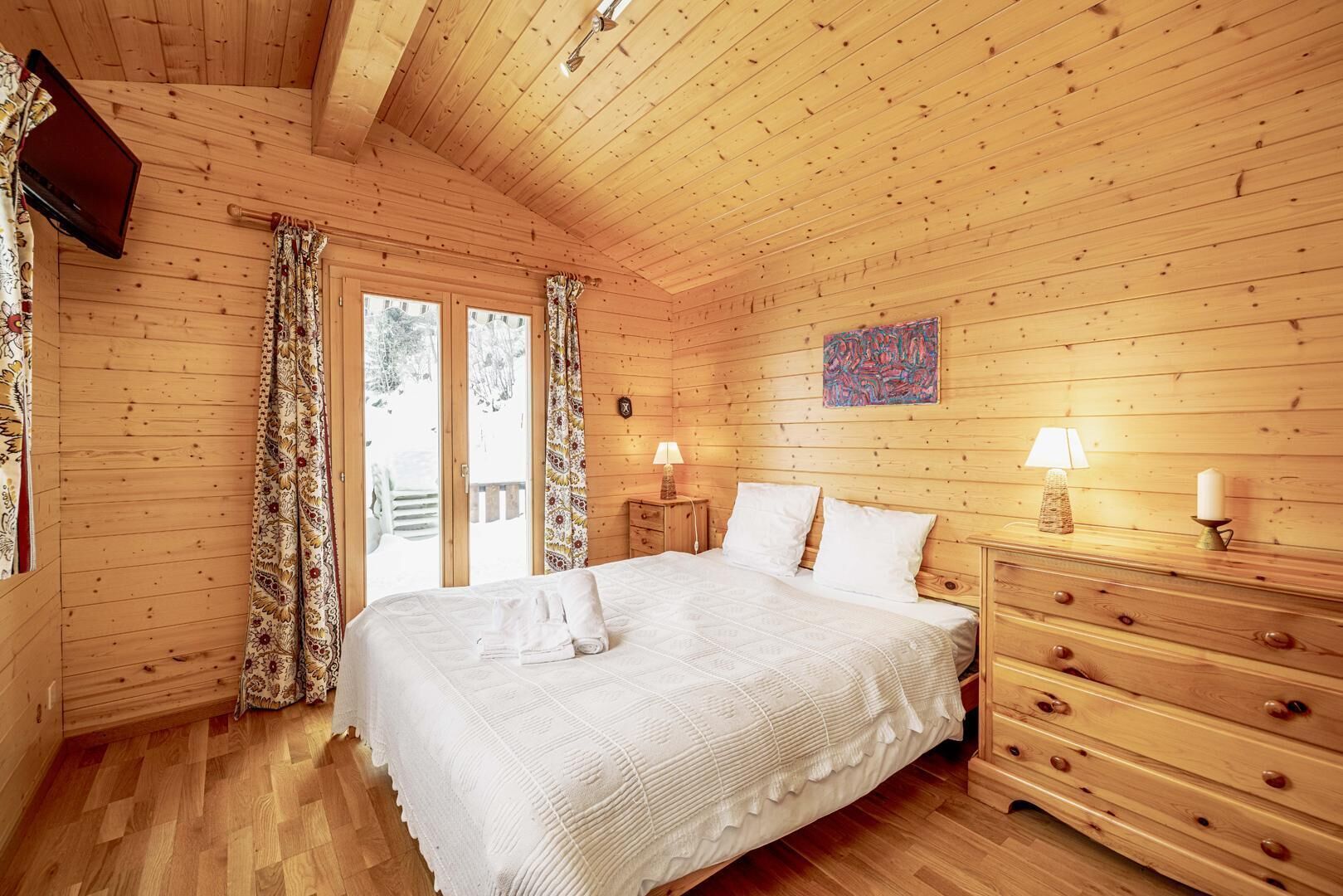 Ett sovrum i en stuga med träpanel, ett stort sängbord, två sängbord med lampor, en byrå och en tavla på väggen.