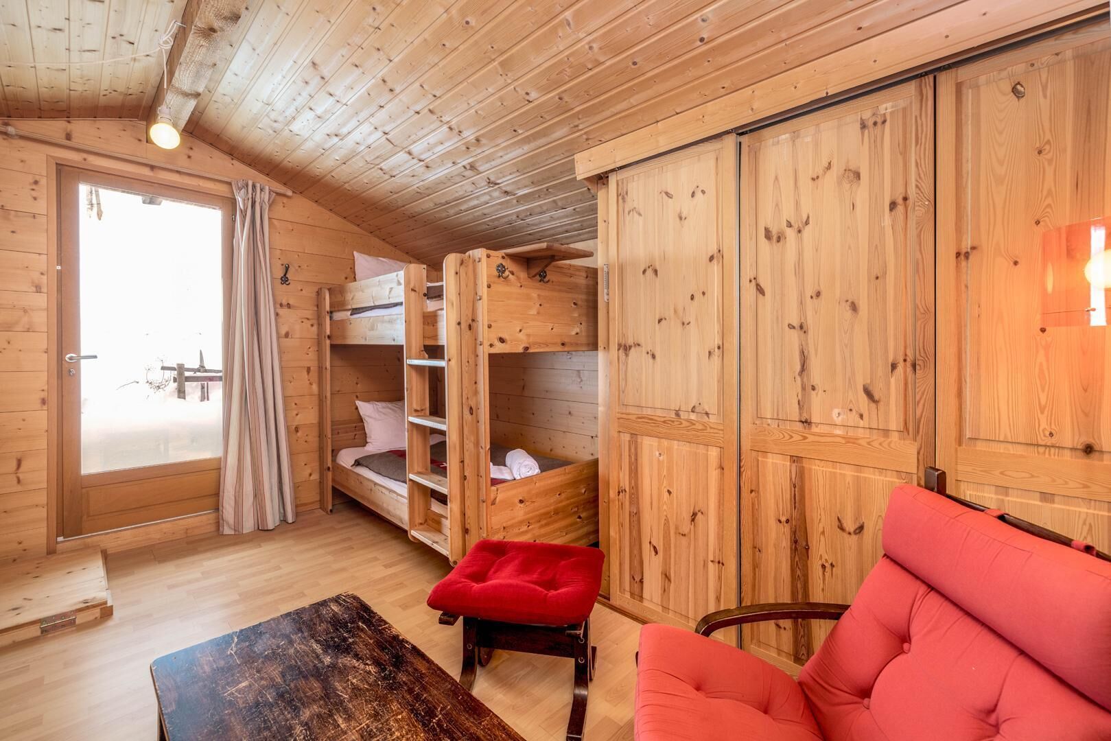 L’intérieur d’une cabane en bois, avec un lit superposé, un fauteuil rouge rembourré et une table en bois.