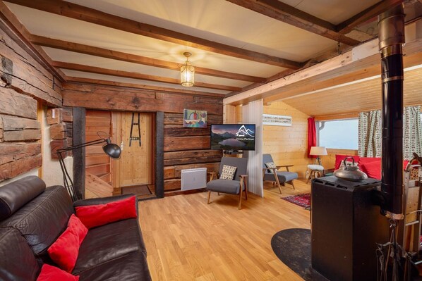 A cozy log cabin interior with a wooden ceiling, a fireplace, a leather sofa, and a television.