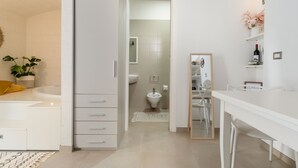 A modern bathroom with a bathtub, toilet, and sink. There is a mirror and a shelf with a bottle. - Almond Tree Suite (Ostuni)