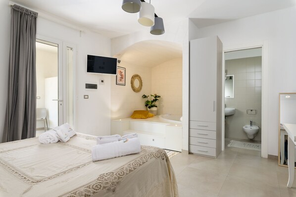A modern hotel room with a large bed, a TV, a small seating area with a plant, and a bathroom with a bidet.