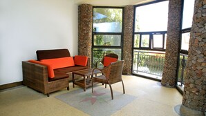 Una habitación con un sofá de mimbre, una mesa de comedor y sillas. Se observa un ventanal con pilares de piedra y un balcón exterior.