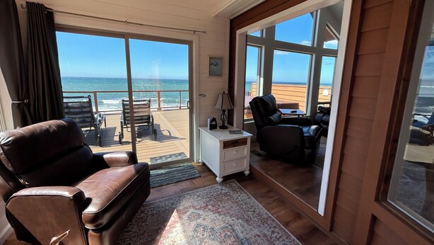 Ein Zimmer mit einer großen SchiebeglasTür, die auf einen Balkon mit Meerblick führt. Zwei Ledersessel und ein kleiner weißer Tisch mit einer Lampe stehen dort.