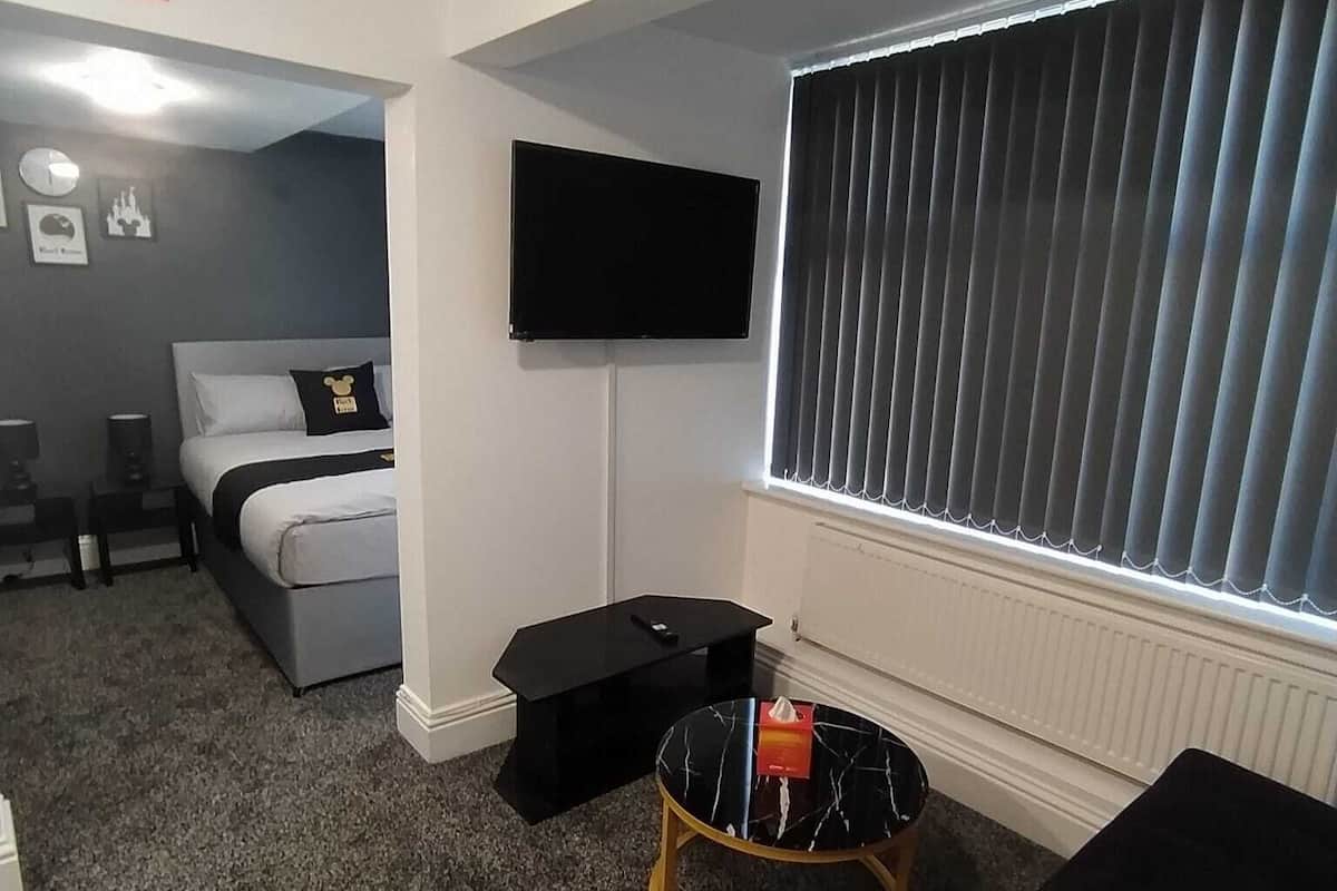 Ein kleines Zimmer mit einem Bett, einem an der Wand befestigten Fernseher, einem kleinen runden Tisch und einem Fenster mit Jalousien.