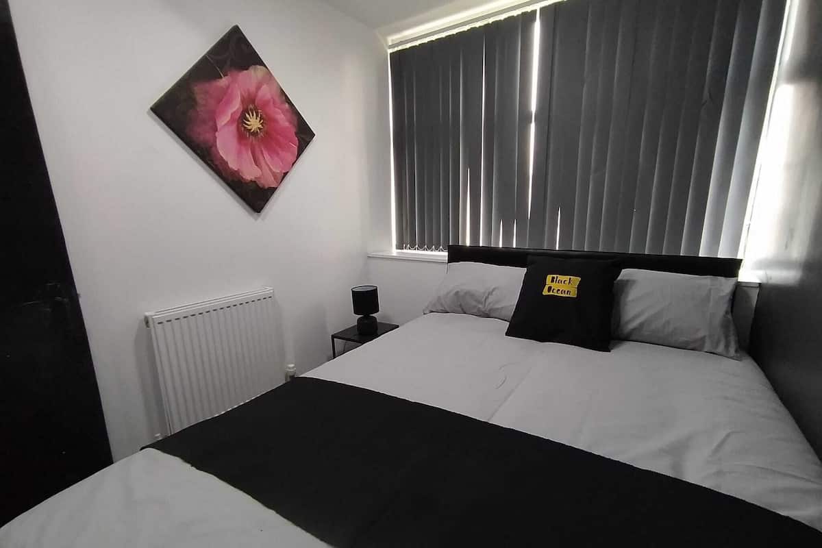 Ein Schlafzimmer mit einem großen Bett, einem Heizkörper, einem Nachttisch mit Lampe, einem Fenster mit Jalousien und einem Wandbild einer Blume.