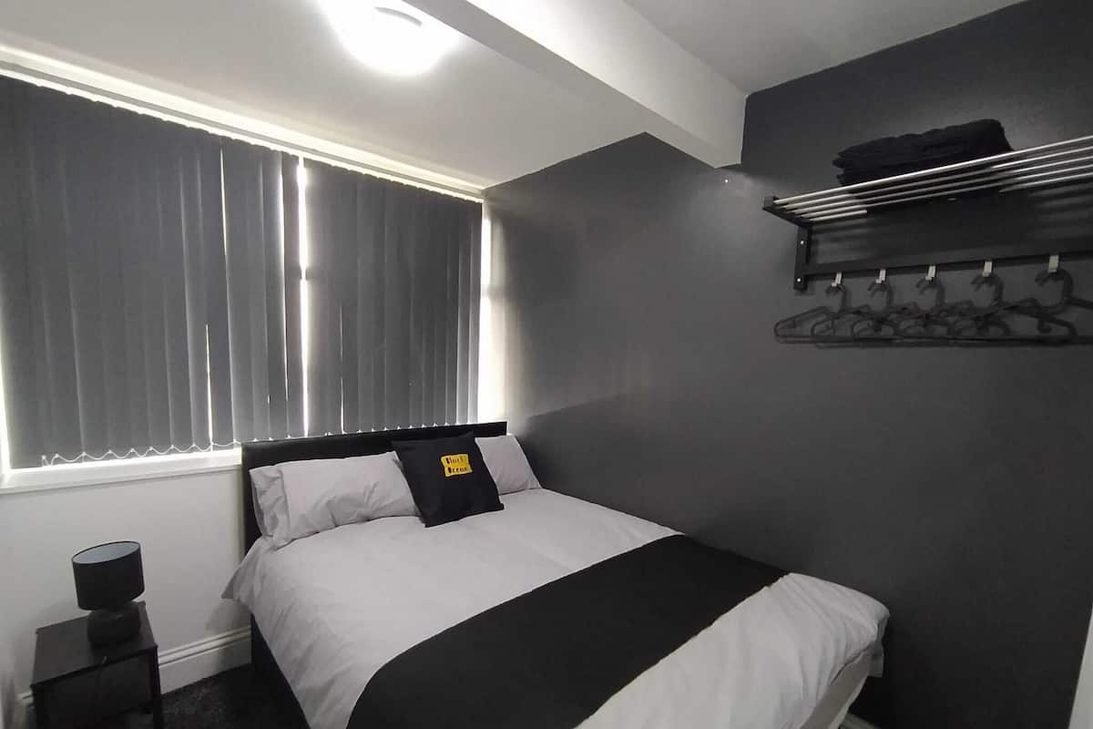 Ein Schlafzimmer mit Bett, Nachttisch, Lampe, einem Fenster mit Vorhängen und einem an der Wand befestigten Kleiderhaken.