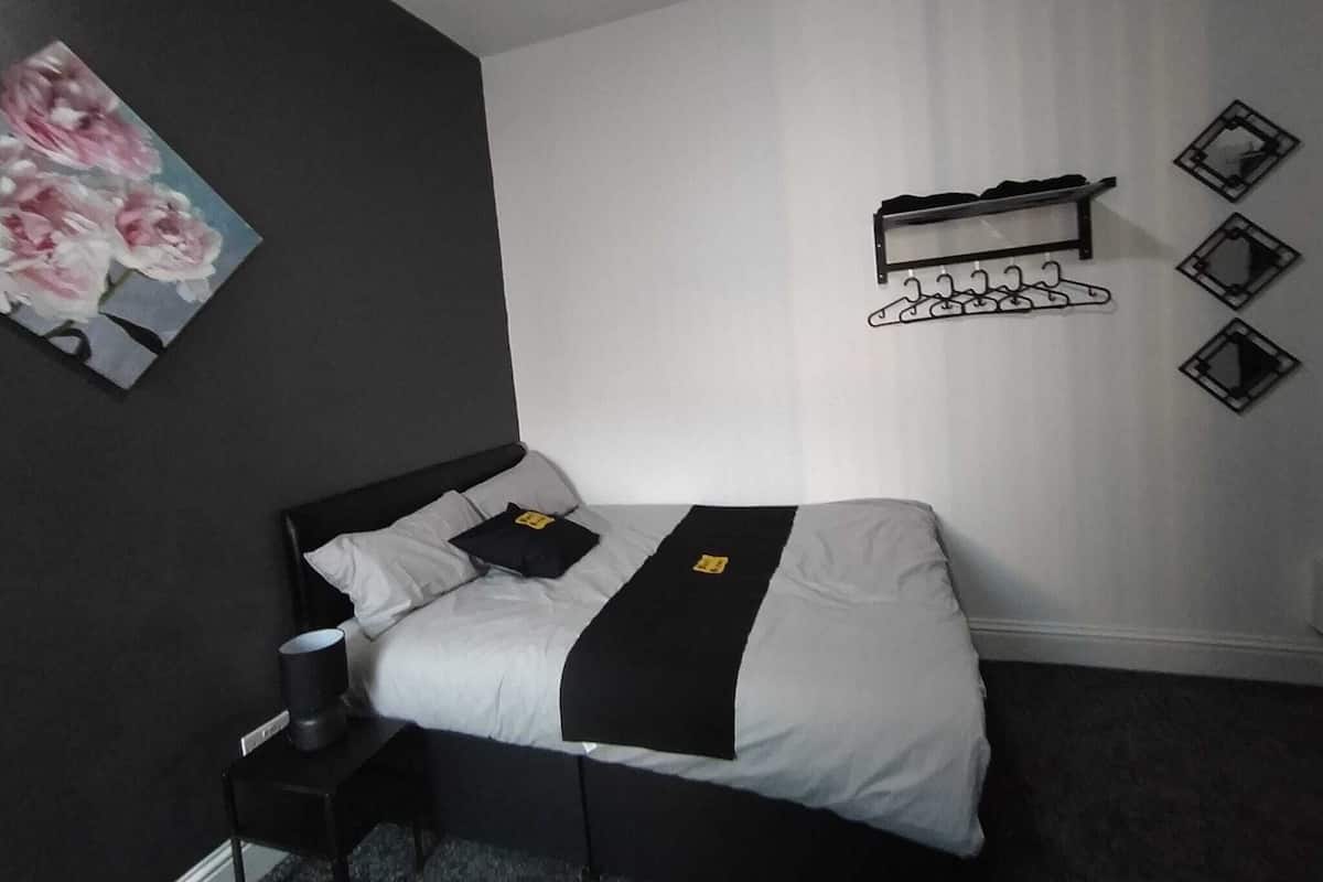 Ein Schlafzimmer mit einem Bett, einem Nachttisch, einem Gemälde und einem an der Wand befestigten Garderobenständer.