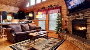 A cozy cabin interior with a fireplace, wooden floors, a leather sofa, a television, and a kitchen area. - Private Deck with Hot Tub & Outdoor TV, Fun Game Loft, Fast Starlink Internet ⭐️ (Sevierville)
