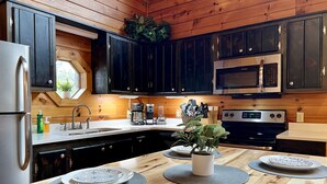 A modern kitchen with dark cabinets, stainless steel appliances, and a wooden dining table set with plates and a potted plant. - Private Deck with Hot Tub & Outdoor TV, Fun Game Loft, Fast Starlink Internet ⭐️ (Sevierville)