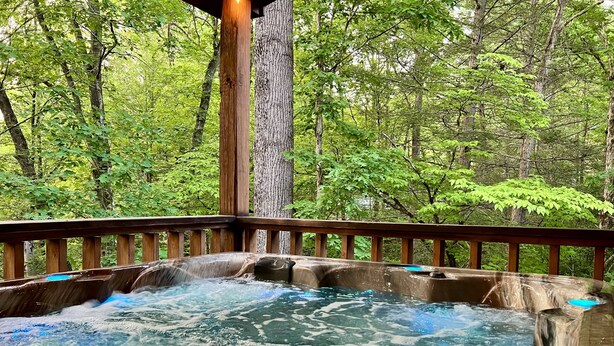 A hot tub with a wooden deck, surrounded by lush greenery. - Private Deck with Hot Tub & Outdoor TV, Fun Game Loft, Fast Starlink Internet ⭐️ (Sevierville)