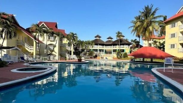 A swimming pool with lounge chairs and palm trees, surrounded by buildings with red roofs. - Bucket List Stays (Ocho Rios)