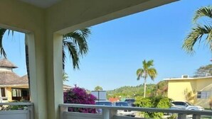 A round glass table with metal legs, two striped chairs, and a view of a residential area with palm trees and parked cars. - Bucket List Stays (Ocho Rios)