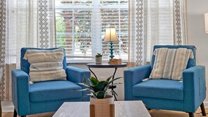 Two blue armchairs with patterned cushions, a small table with a plant, and a window with blinds and a lamp.