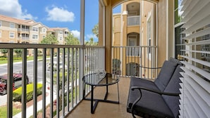 A balcony with a round glass table and two chairs, overlooking a street with parked cars and a residential building in the background.