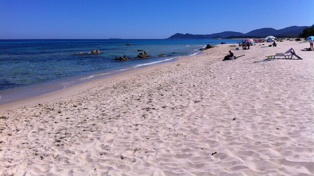 A sandy beach with clear blue water, rocks, and mountains in the distance.