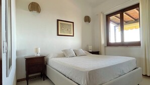 A bedroom with a large bed, a bedside table, a wall-mounted lamp, a framed picture, and a window with curtains.