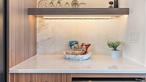 Eine Küche mit Marmor-Spritzschutz, einer weißen Arbeitsplatte, einer Schüssel mit Snacks, einem Topf mit Pflanze und einem Regal mit Gläsern und einem schwarzen Behälter.