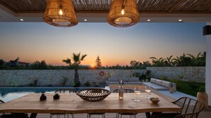 Un espace extérieur aménagé pour les repas, avec un mur en pierre, une piscine et une vue sur le coucher de soleil.