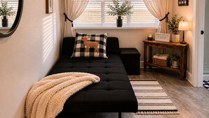 Una camera da letto moderna con un divano letto nero, una soffice coperta beige, un comodino con una lampada e una finestra con tende leggere.