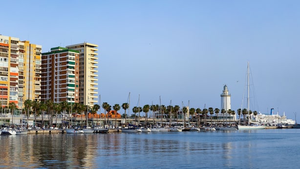 Uma marina com barcos, um farol e edifícios altos ao fundo.