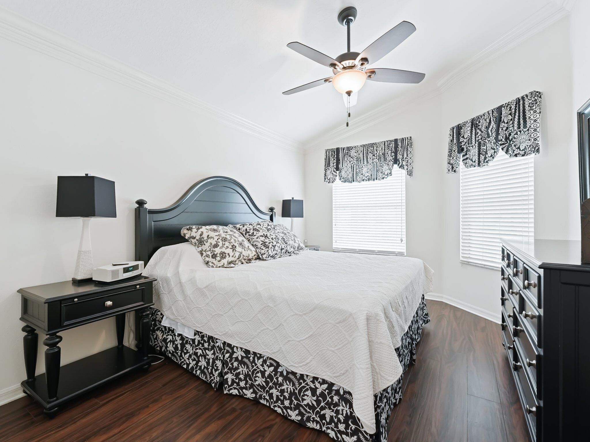 A bedroom with a bed, a nightstand, a dresser, a ceiling fan, and patterned curtains.
