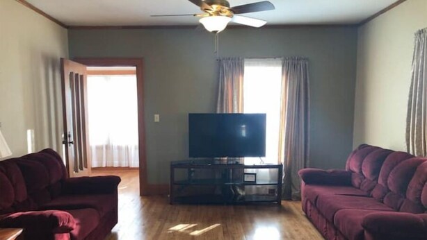 A living room with two dark sofas, a wooden floor, a ceiling fan, a TV on a stand, and a door leading to another room.