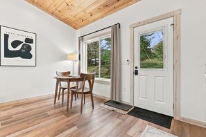 A room with a wooden floor, a white door, a window with curtains, a framed artwork on the wall, and a table with chairs. - Onyx - Near Hocking Hills (Rockbridge)