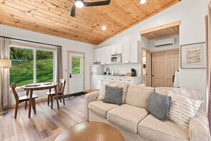 A modern living room with a wooden ceiling, a beige sofa with striped pillows, a dining table with chairs, and a kitchen area with white cabinets and appliances. - Onyx - Near Hocking Hills (Rockbridge)