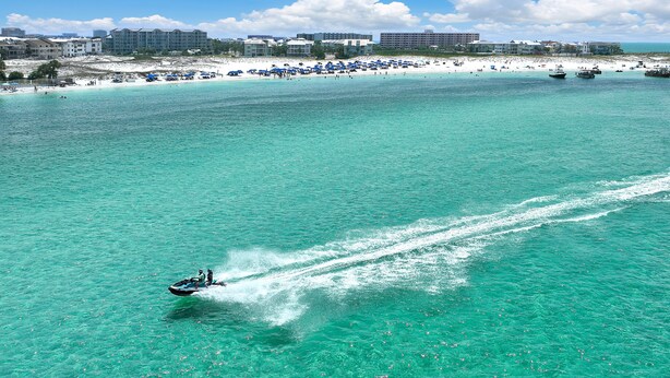 A beach with clear turquoise water, a jet ski, and a distant shoreline with buildings. - 4BR Spacious Beach Home with Private Pool+ Studio Apartment | Southern Nights (Destin)