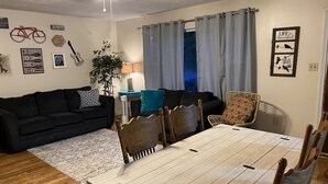 Een woonkamer met een zwarte bank, een eettafel met stoelen, een plafondventilator en diverse wanddecoraties.