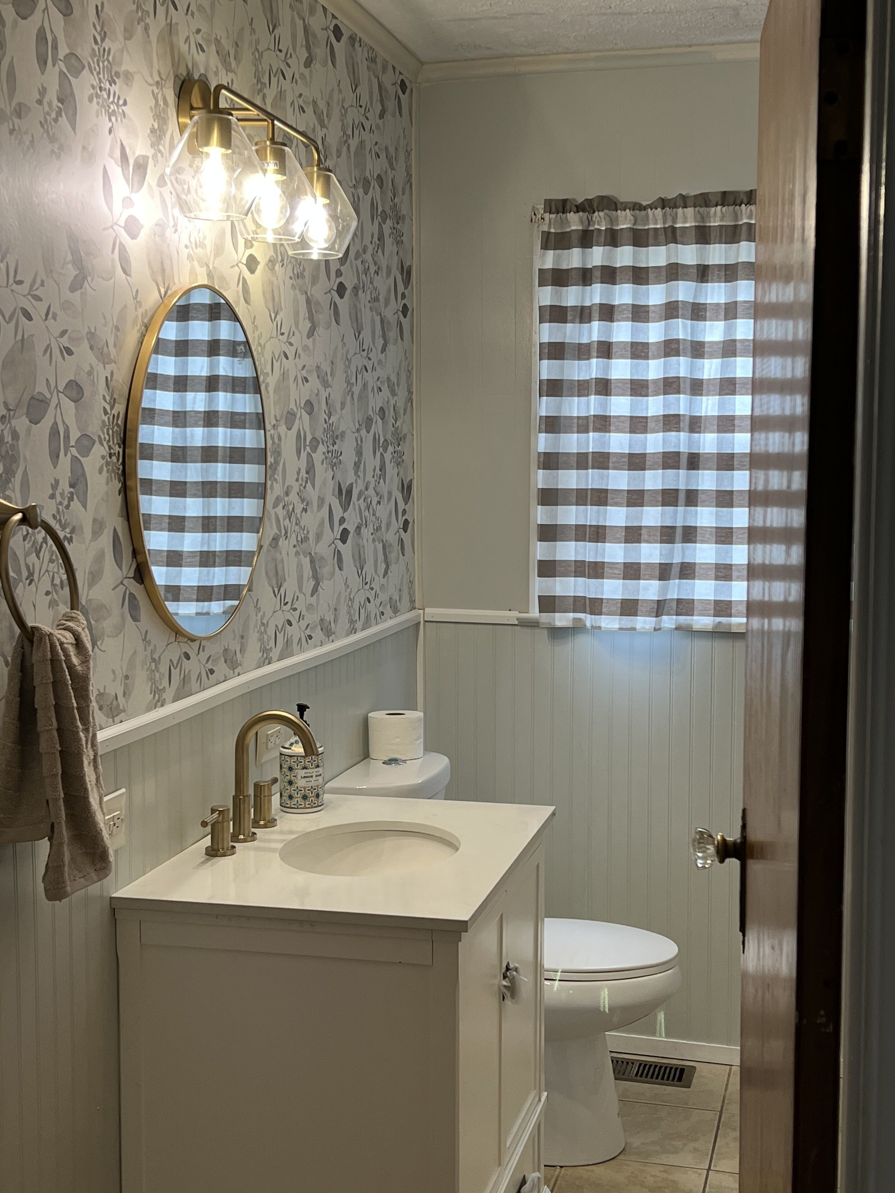 Une salle de bain avec un meuble-lavabo, des toilettes et une fenêtre ornée d’un rideau à carreaux.