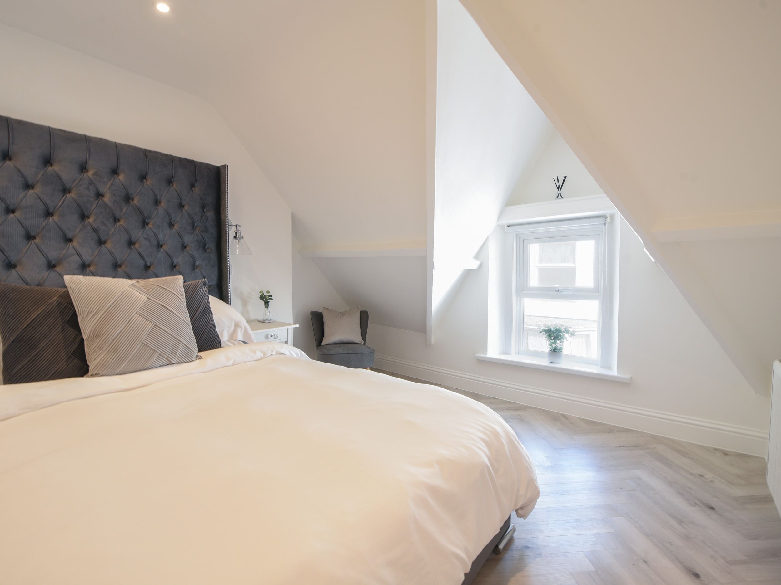 A bedroom with a large bed, a tufted headboard, a small table with a lamp, a chair, and a window with a view.