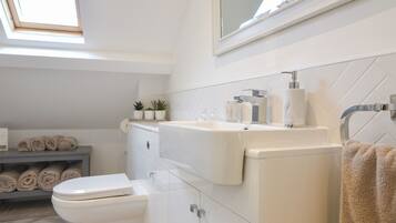 A modern bathroom with a toilet, sink, and shower area. There is a skylight above the toilet, and towels are neatly rolled on a shelf.