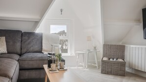 Ett modernt sovrum i loftstil med en grå soffa, ett träbord, ett litet vitt bord, en korg av rotting och utsikt över naturen genom ett fönster.