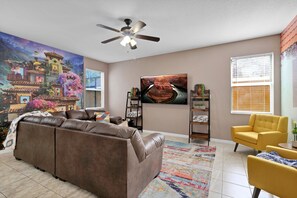 A living room with a brown sofa, a yellow armchair, a ceiling fan, a TV, and a large mural on the wall.